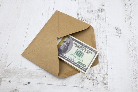 A brown envelope with a bundle of 100 dollars lies on a white wooden table