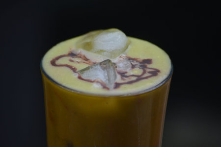 Tall glass of iced mango drink with visible syrup streaks and ice cubes isolated against dark...