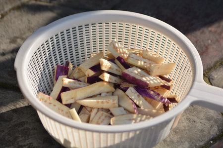 Fresh raw eggplant cut into sticks and chunks resting in a white strainer colander with natural sunlight and shadow pattern. Food preparation concept for cooking healthy vegetable mealの写真素材