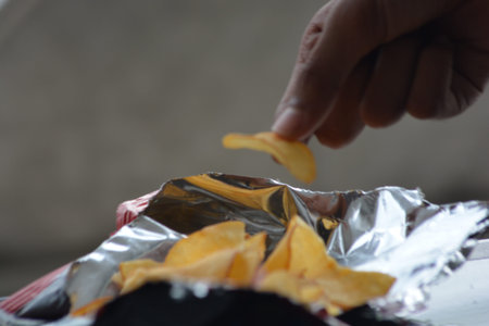 Human hand taking a single potato chip from an open foil package concept of snacking choice temptation sharing and lifestyle moments useful for diet and habit articlesの写真素材