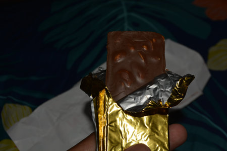 Close up of a chocolate bar partially wrapped in silver and gold foil being held by a hand, suitable...
