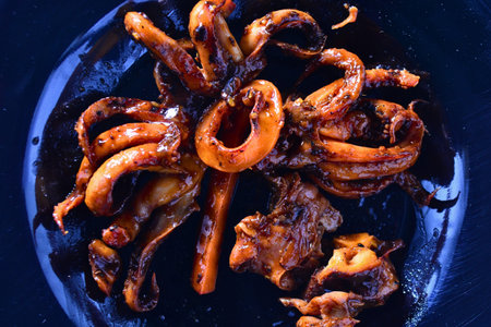 Close up macro of a glossy caramelized calamari ring centered among curled squid tentacles on a dark surface. Rich color and texture ideal for menu headers cooking blogs seafood advertising and recipe illustrations.の写真素材