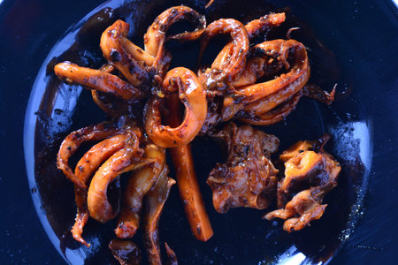 Top view tight crop of grilled squid with glossy sweet spicy glaze and curled tentacles on a dark plate. Strong textures and rich orange brown tones suitable for food delivery banners recipe cards and culinary blogs.の写真素材