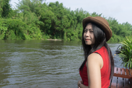 Happy Asian girl red shirt and brown hat on the river in nature background, Relax time on holiday concept travel., Thailand river kwai and typical landscape kanchanaburi., with copy space for textの写真素材