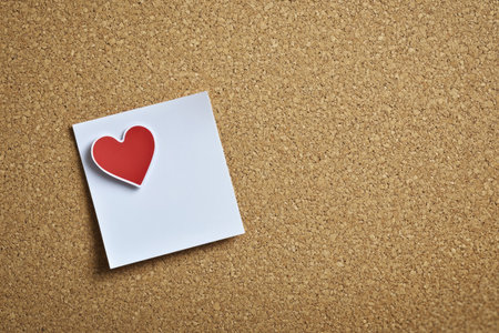 A vibrant red heart sticker placed on a blank white sticky note, pinned to a textured cork board. Copy space available for messages.の素材