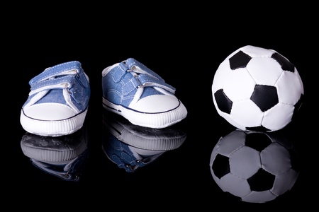 pair of small baby shoes next to football ball, footballer baby or baby footballer concept, studio shoot isolated on black with reflectionの写真素材
