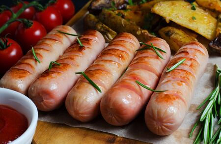Grilled sausages on a wooden chopping Board. Fried potatoes, rosemary, tomatoes, tomato ketchup. Unhealthy diet.の写真素材