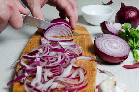 A man cuts red onions on a wooden chopping Board with a kitchen knife. Hands close up. Preparing...
