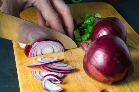 A man cuts red onions on a wooden chopping Board with a kitchen knife. Hands close up. Preparing food. Side view.の写真素材