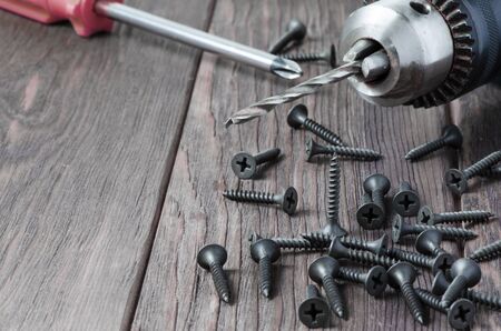 Metal screws and an electric drill on a wooden background. Tool for fixing and repairing.