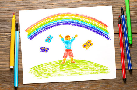 The boy rejoices in the rainbow. Childrens drawing on white paper. Wooden table with markersの写真素材