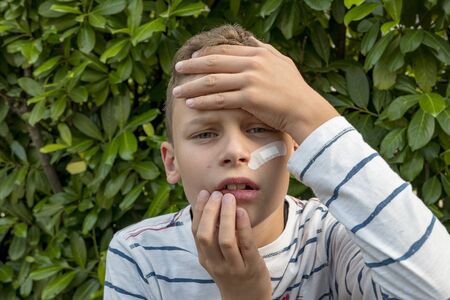 Boy with plaster on his face in front of a hedge looks sadの写真素材