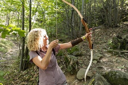 blonde woman shooting with a bow and arrow in the forest