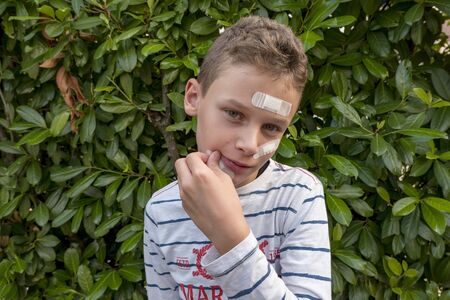 Boy with plaster on his face in front of a hedge looks sadの写真素材