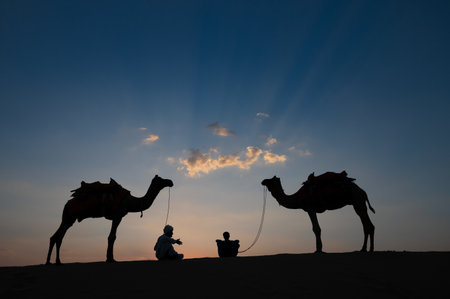 Thar desert, Rajasthan, India- 15.10.2019 : Silhouette of two cameleers and their camels at sand dunes. Cloud with setting sun, sky background. Cameleers make a living out of camel riding by tourists.のeditorial素材