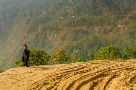 Sikkim, India - 22nd March 2004 : A school boy in uniform, waiting for his school bus in morning, beside curved soil road with Himalayan mountains in the background.の写真素材
