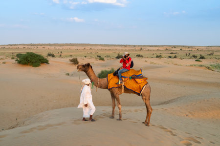 Female tourist taking picture while riding camel, Camelus dromedarius, at sand dunes of Thar desert, Rajasthan, India. Camel riding is a favourite activity amongst all tourists visiting here.のeditorial素材