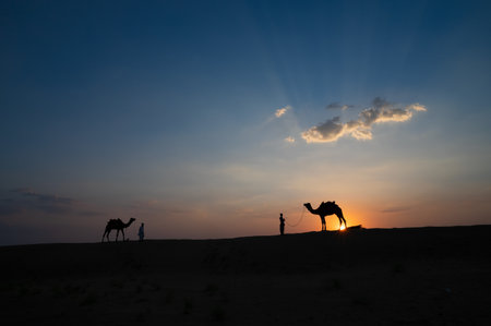 Silhouette of two cameleers and their camels at sand dunes of Thar desert, Rajasthan, India. Cloud with setting sun, sky in the background. Cameleers make a living out of camel riding by tourists.のeditorial素材