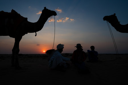 Silhouette of two cameleers with a female tourist. Camels at sand dunes of Thar desert, Rajasthan, India. Sunset with blue sky in the background. Cameleers use camel riding by tourists, for a living.のeditorial素材