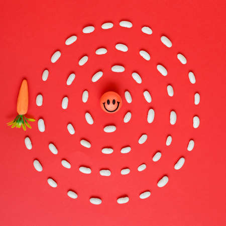 Smiley face ball and artificial carrot conected with bean grains spiral. Flat lay minimal concept on red backgroundの写真素材