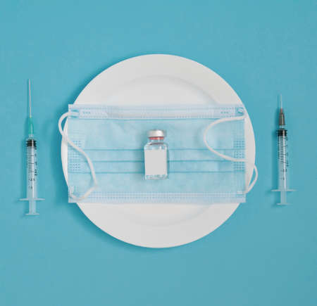 The vial and face mask lay down on a plate, with two syringes by the side. Minimal flat lay pandemic...