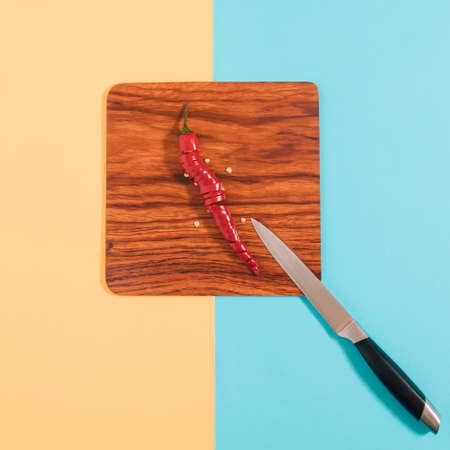 Kitchen knife and sliced red chili peppepr on a brown wooden plate, lay down on a pastel yellow and...