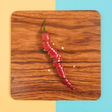 Sliced red chili peppepr on a brown wooden plate, lay down on a pastel yellow and blue background,...