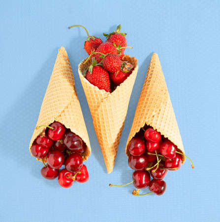 Ice cream cones filled with ripe strawberries and cherries lie down on a blue background. Square...