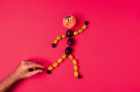 A human figure made of grape grains, plums and peach runs away from the male hand. Minimalistic creative funny autumn concept, flat lay composition on pastel red backgroundの写真素材