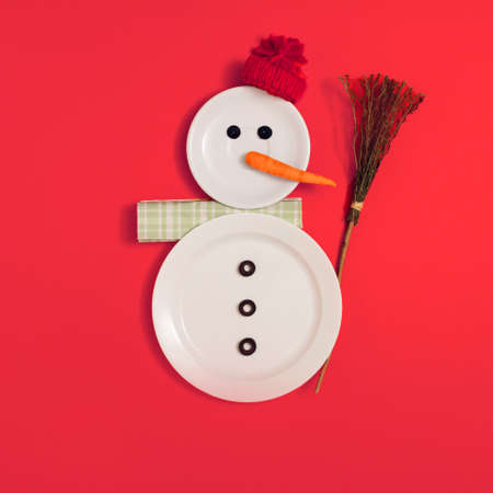 Snowman with broom and hat, made of ceramic plates, napkin, sliced olives and carrot. Square flat lay composition on red background, minimal creative funny Christmas or New Year decorative conceptの写真素材