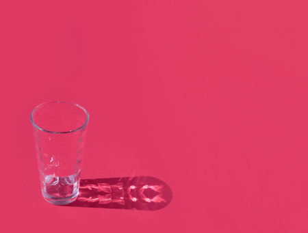 Emtpty water glass with a fancy shadow on pastel red background. Minimal horzontal composition with copy spaceの写真素材