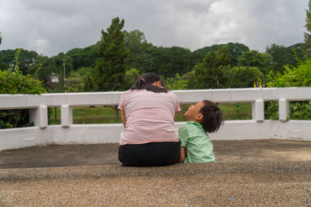 Back view of loving Asian mother hug her kids sitting on bridge, caring black mom embrace child, relaxing looking to black cloud, parent comfort child caressing.の写真素材