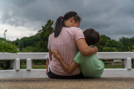 Back view of loving Asian mother hug her kids sitting on bridge, caring black mom embrace child, relaxing looking to black cloud, parent comfort child caressing.の写真素材
