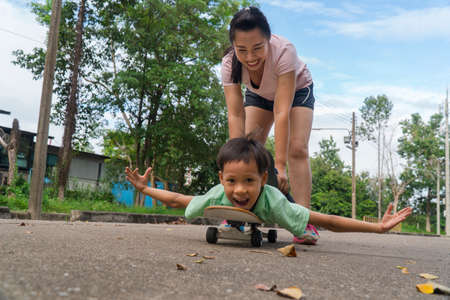 Young asian mother playing with her son pushing forward boy riding on skateboard in public road. Kids and entertainment. Childhood and sports concept.の写真素材