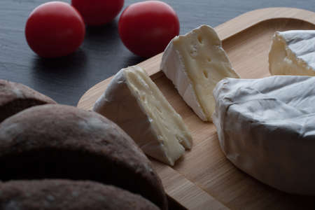 Fresh French Brie cheese and a one slice on a bamboo board with cherry tomatoes, bread on the black plate.の写真素材