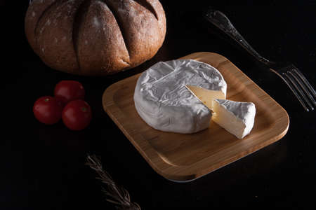 Fresh French Brie cheese and a one slice on a bamboo board with cherry tomatoes, bread and fork on the black plate.の写真素材