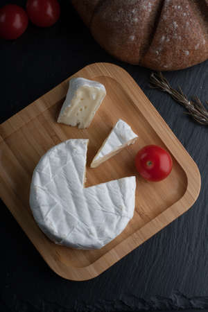 Fresh French Brie cheese and a one slice on a bamboo board with cherry tomatoes, bread on the black plate. top view.の写真素材