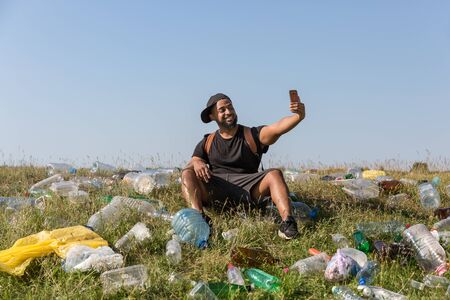 smiling young man with a phone sitting on a green slope strewn with plastic bottlesの写真素材