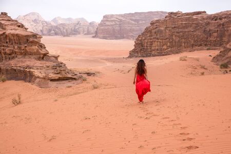a girl with long hair in a red long dress goes into the desert. Dress develops in the wind. Traces remain in the sandの写真素材