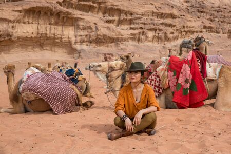 A camel driver in a cowboy hat and boots sits on the sand among the herd against a background of...