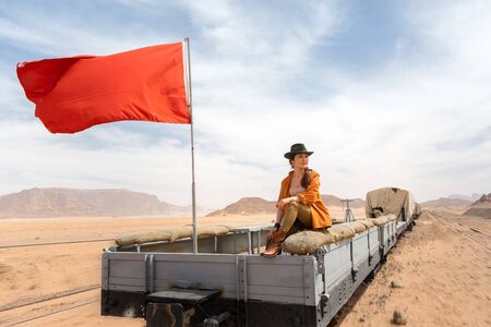 A girl in a cowboy hat rides on a wooden carriage of an old train in the Wadi Rum desert. A red flag...