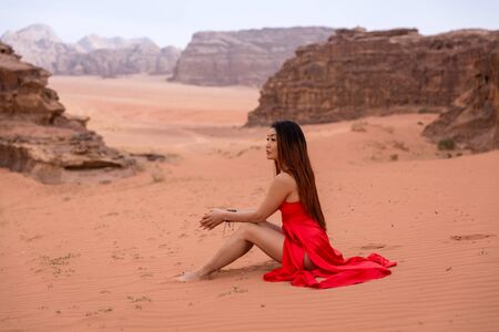 A cute Asian girl with long hair in a red long dress sits on the sand and looks into the distance. In the background the desert and mountains. Shot in Jordan Wadi Rum Desertの写真素材