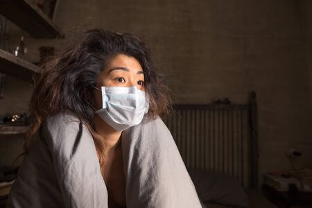 A girl in a medical mask sits on a bed wrapped in a blanket and looks out the windowの写真素材