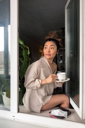 A girl in a mans shirt with a cup of coffee sits on a windowsill and looks out into the streetの写真素材