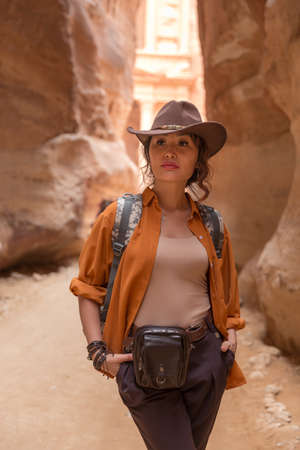A beautiful girl with a backpack in a cowboy hat and an orange shirt stands at the bottom of the canyon and looks aheadの写真素材