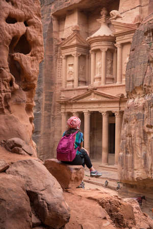 Girl tourist with a backpack sits on a stone and looks at El-khazneh in the Siq canyonの写真素材