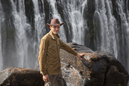 a man in a cowboy hat and a hakki shirt stands on a rock with a waterfall in the backgroundの写真素材