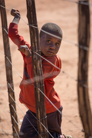 Namibia, Africa, Himba village, July 18, 2019: a black boy stands behind a barbed wire fence in torn clothes.のeditorial素材