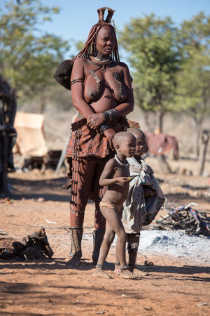 Namibia, July 18, 2019. A Himba woman stands with her children by the fire in traditional topless dress.のeditorial素材