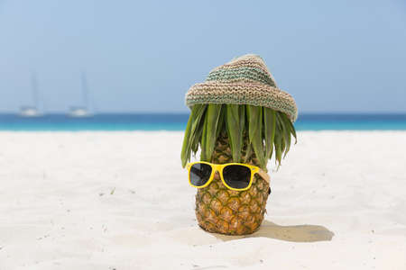 A ripe and juicy mustachioed pineapple in a hippie hat and yellow sunglasses stands on the beach, on white sand, in the background the azure sea and sailboatsの写真素材